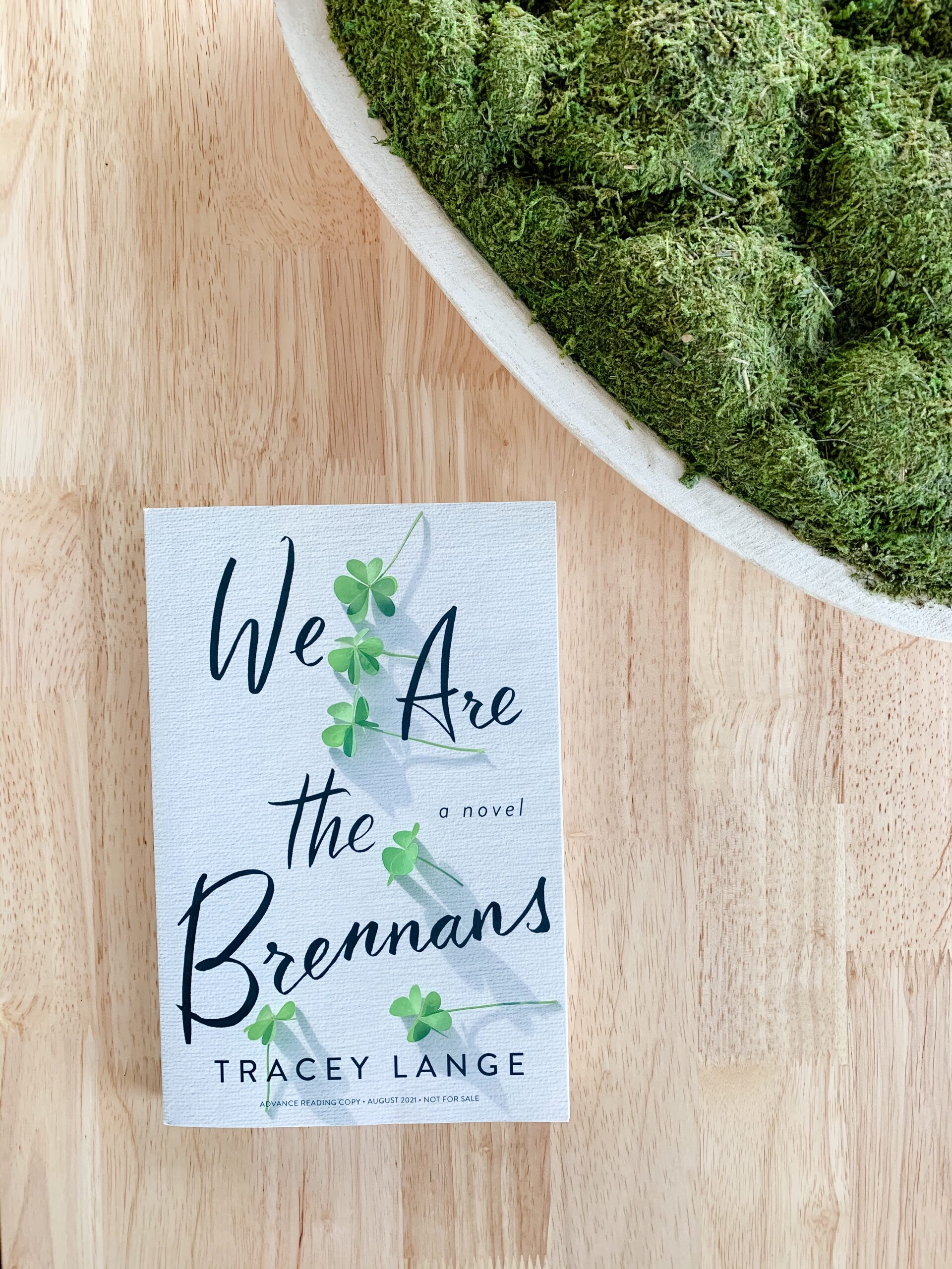 image of We Are the Brennans novel by Tracey Lange. Book is set on oak table with green moss. 